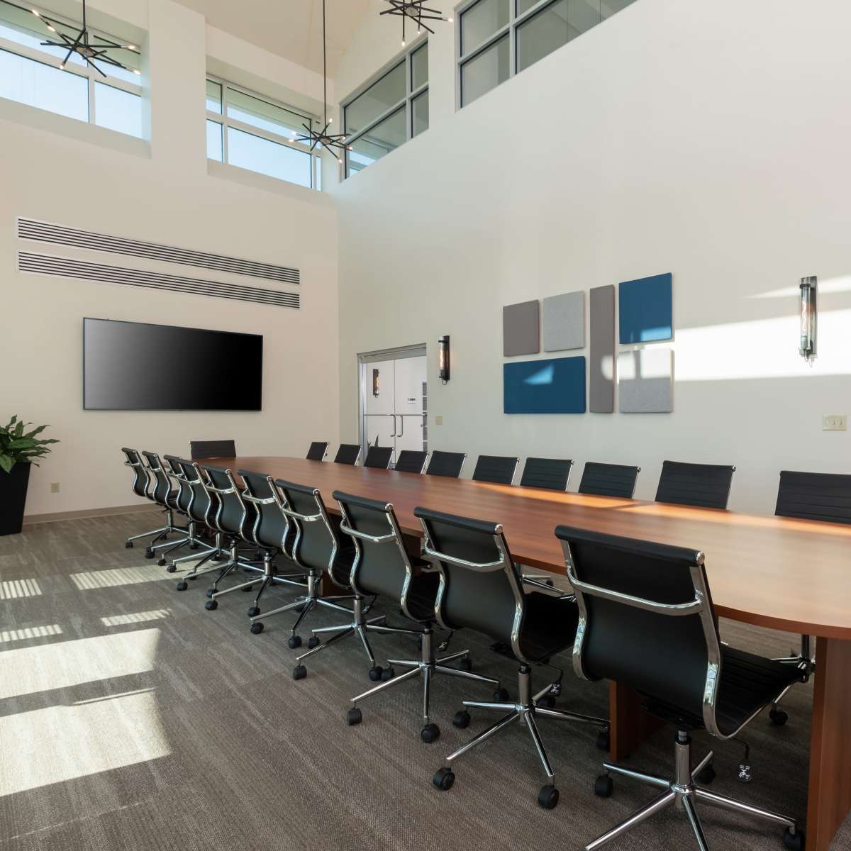 A large conference room features a long wooden table surrounded by ergonomic black chairs, with a large screen mounted on the wall and bright natural light streaming in through tall windows.