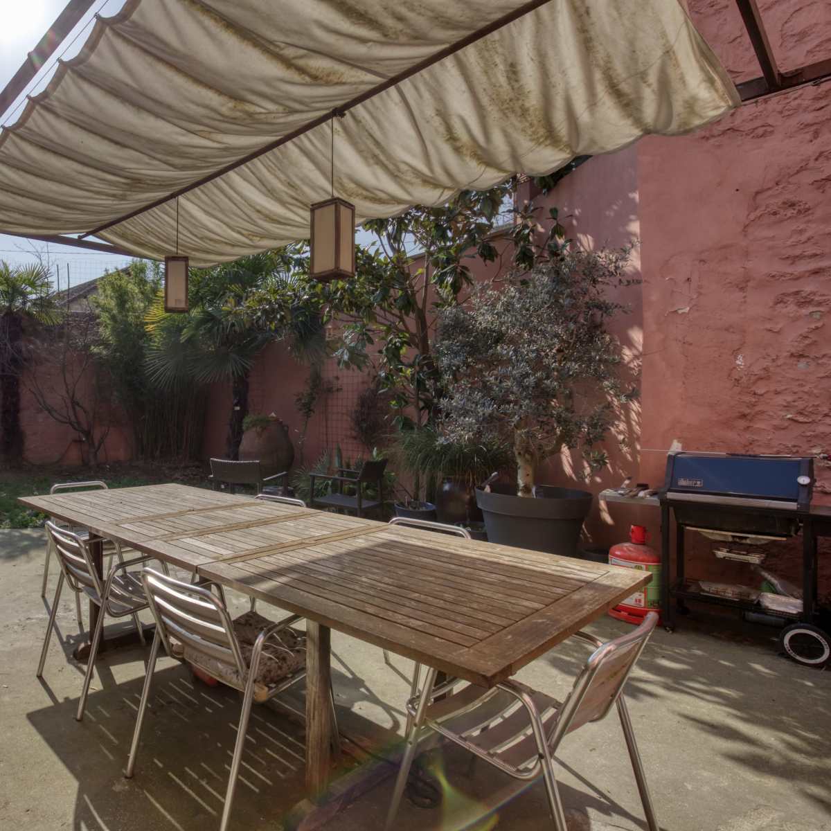 An outdoor patio area with a large wooden table, metallic chairs, a grill, and various plants surrounded by pink walls.