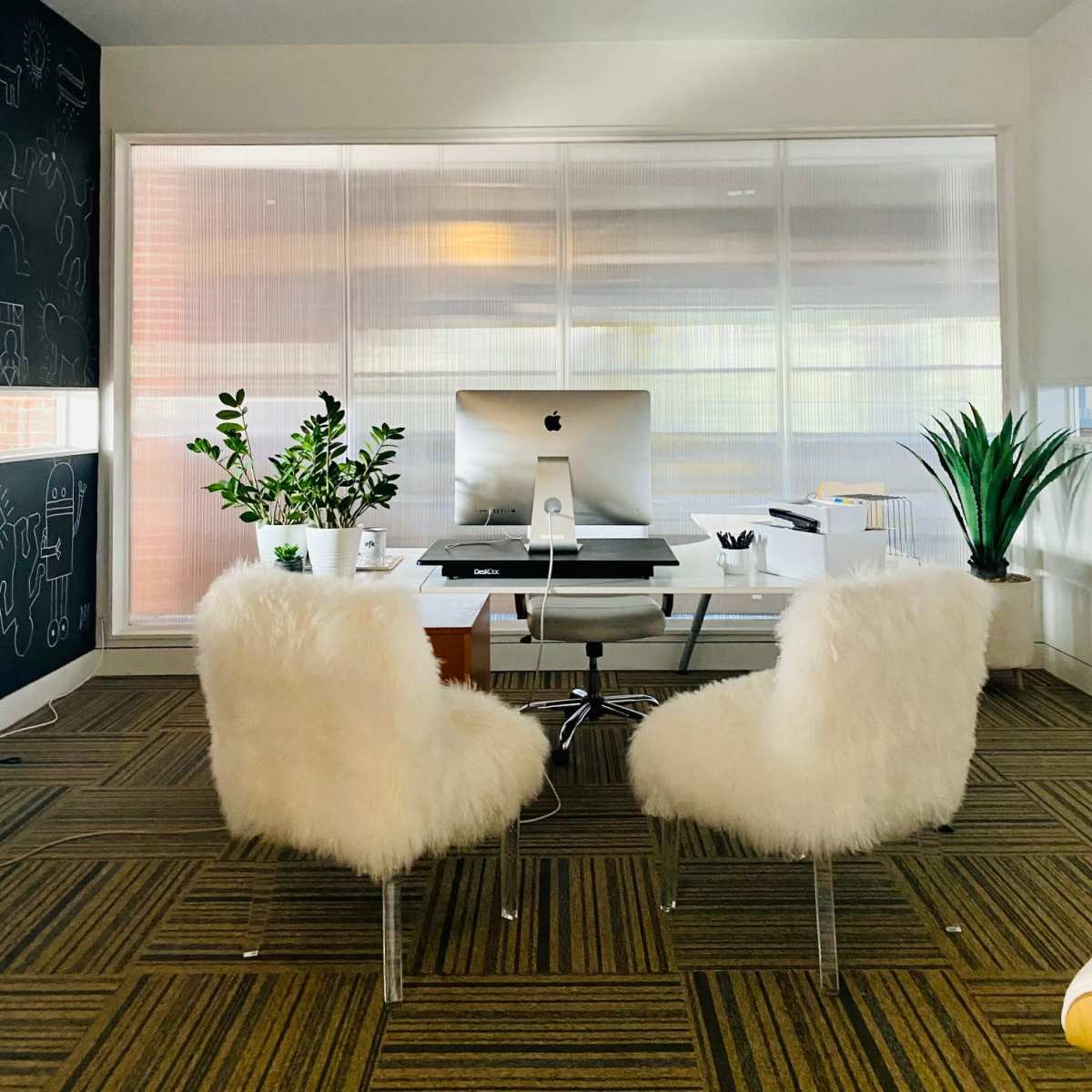 A modern office space features a white desk with an iMac, two fluffy chairs, and potted plants beside large windows.
