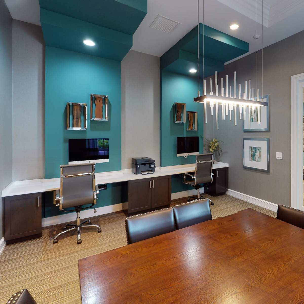 A modern office space featuring a wooden conference table, two computer workstations against turquoise walls, and a glass door leading to a room with plants.