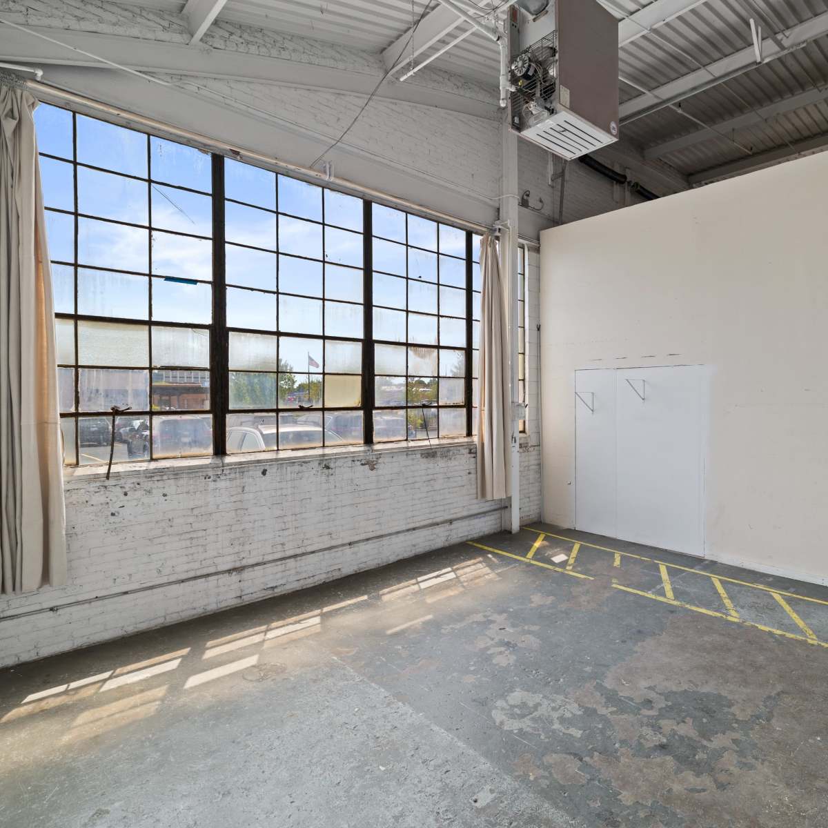 A spacious, empty room features large industrial windows, a blank wall, and marked flooring with yellow lines.