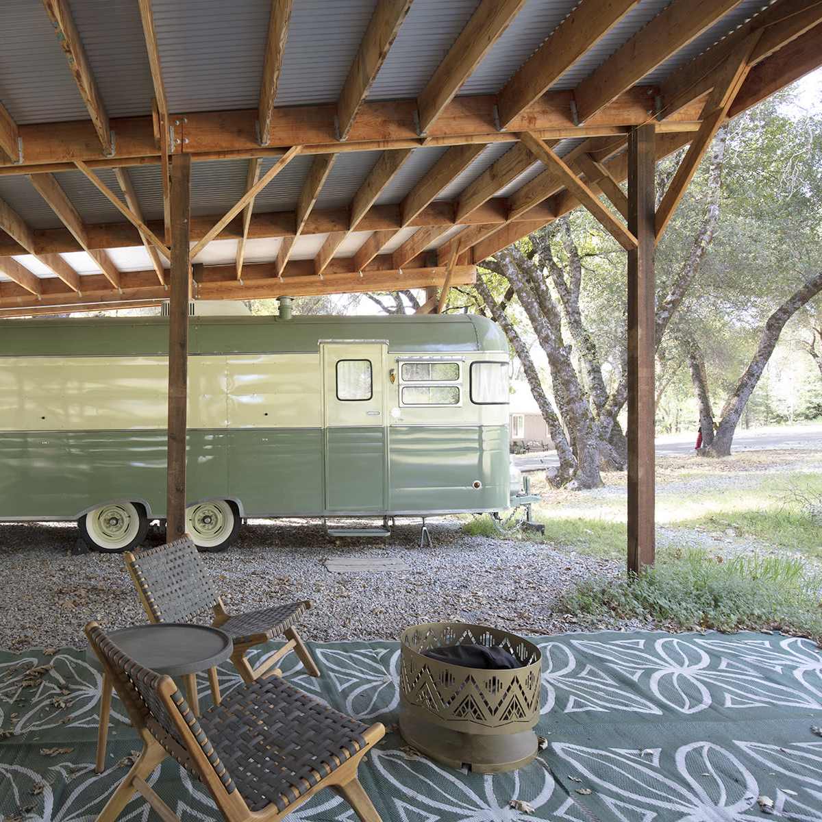 A vintage green and white travel trailer is parked under a wooden pavilion surrounded by trees on a gravel area.