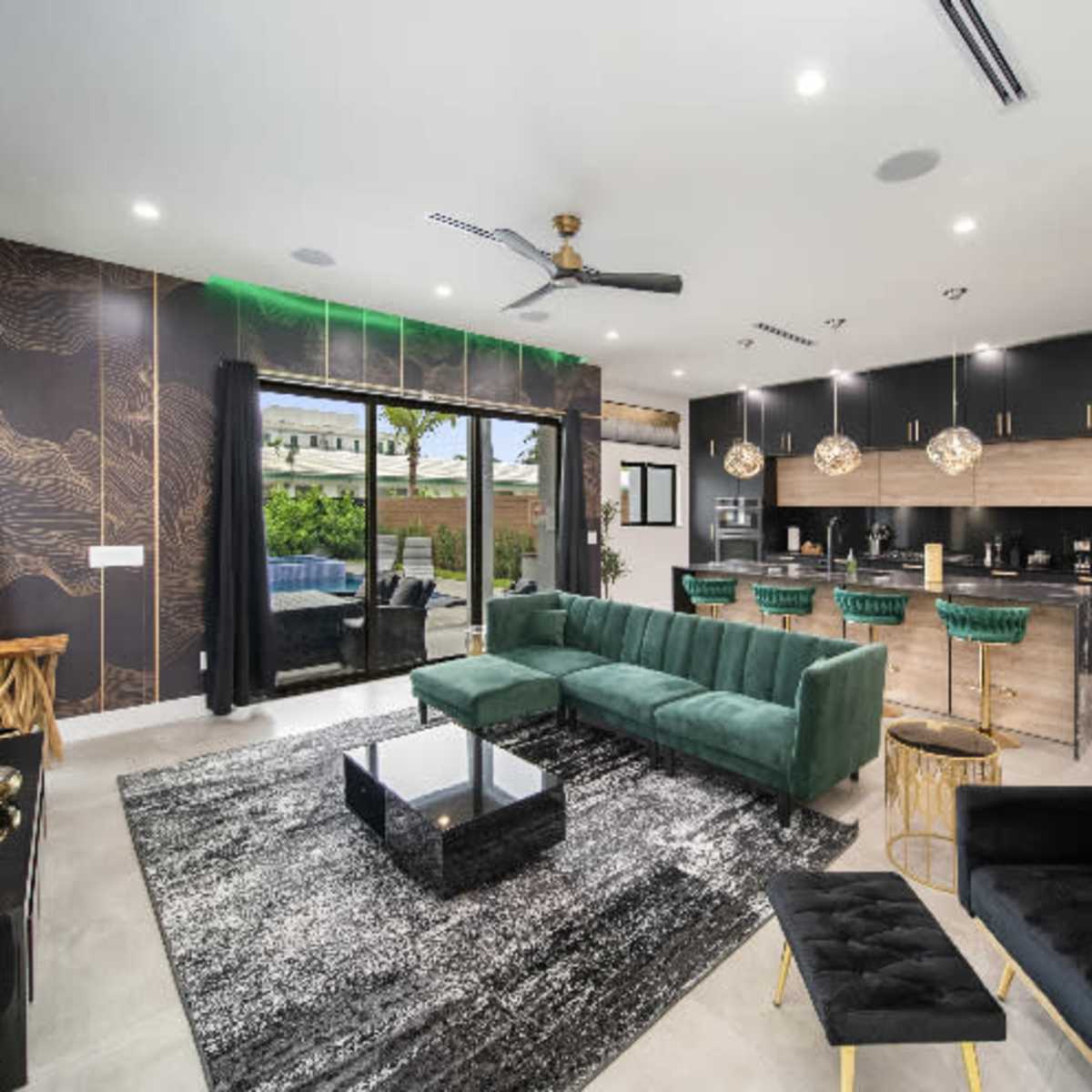 A modern living room features a green velvet sectional, a black coffee table, and a sleek kitchen with dark cabinetry and gold accents.