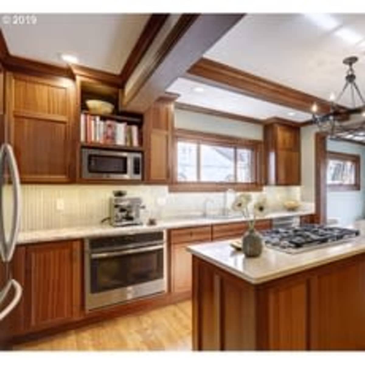 A modern kitchen features wooden cabinetry, stainless steel appliances, a large island with a gas range, and a dining area with large windows.