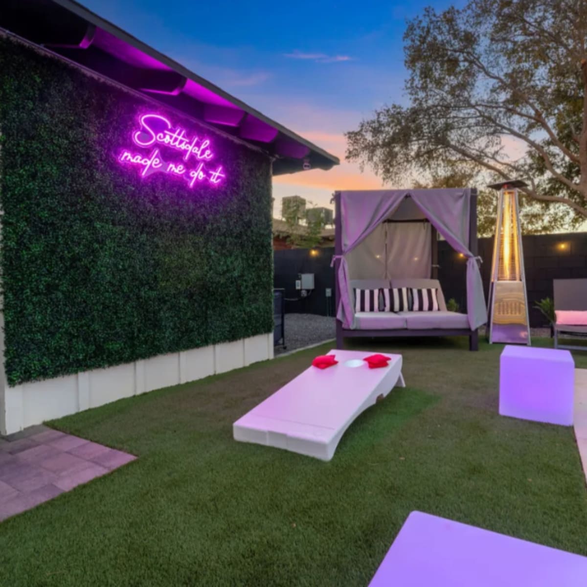 A modern outdoor space features a swimming pool, a lounging area with pink chairs, a canopy bed, and neon signage on a green wall.