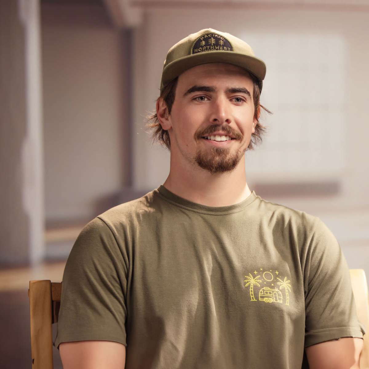 A man wearing a green cap and shirt sits on a chair in a brightly lit room with a wooden floor.