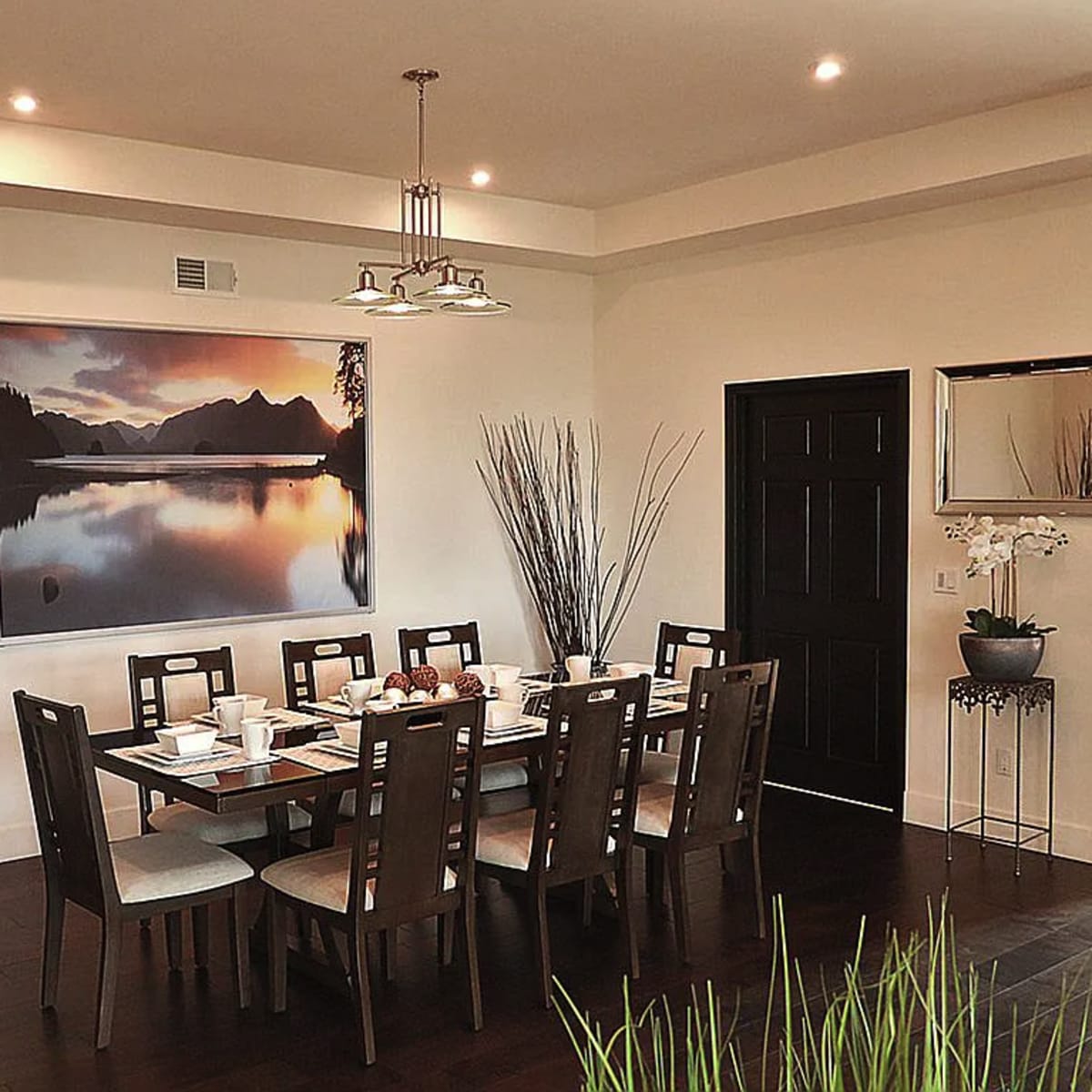 A dining room features a large table set for a meal, surrounded by wooden chairs, with a scenic mountain landscape photograph hanging on the wall.