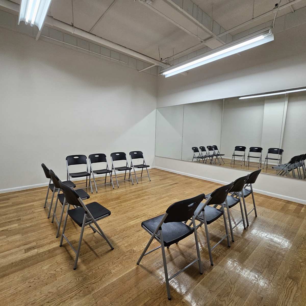 A minimalistic room with wooden flooring and two rows of black chairs arranged in a semicircle facing a mirror.