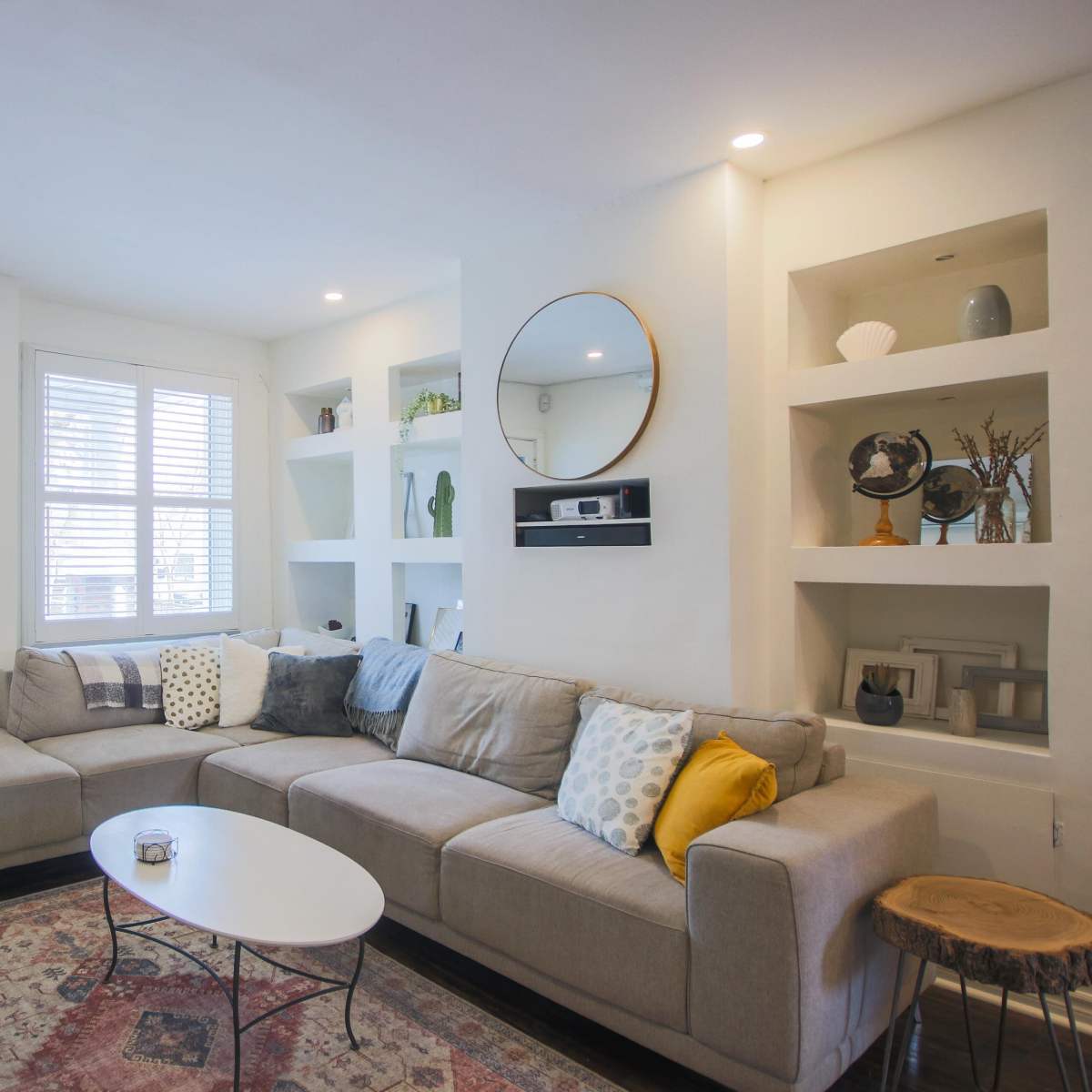 A modern living room features a sectional sofa, a round mirror, and built-in shelves, with a small coffee table and a decorative rug.