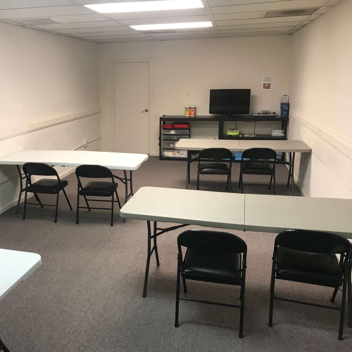 A bare room with several folding tables and chairs arranged, featuring a small television and storage area.