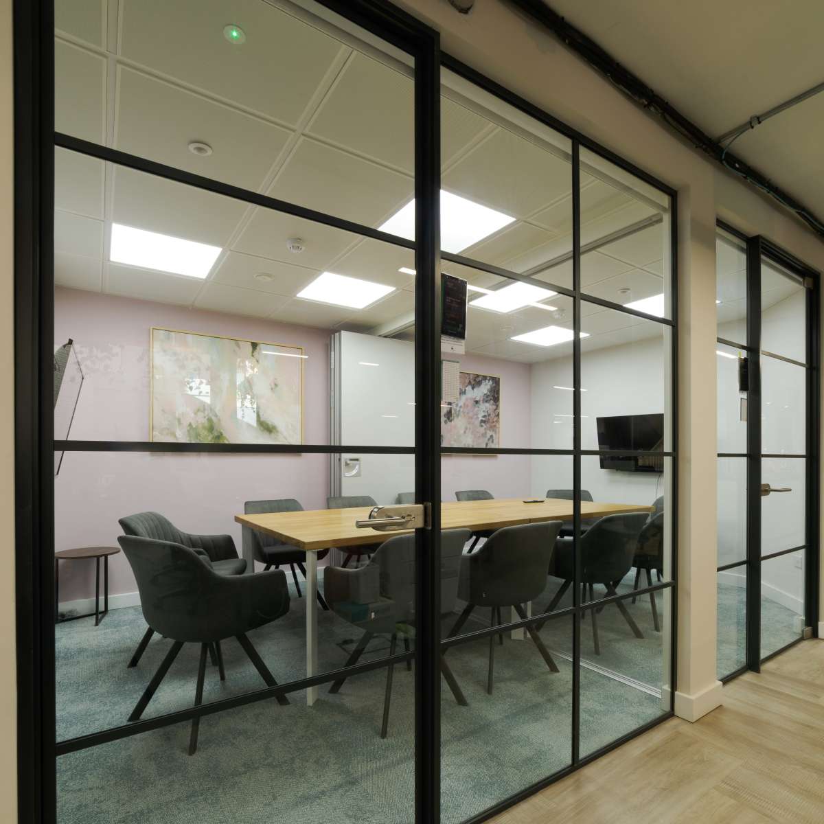 A modern conference room with a large wooden table and gray chairs, partially enclosed by glass walls.