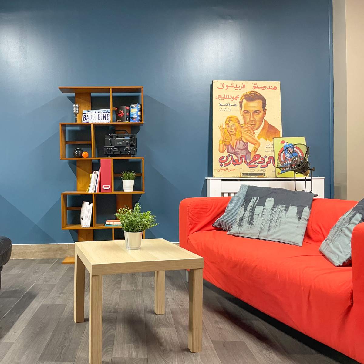 A living room with a black leather couch, a red fabric couch, a wooden coffee table, a bookshelf with various items, and a wall adorned with a vintage poster and a framed photograph.