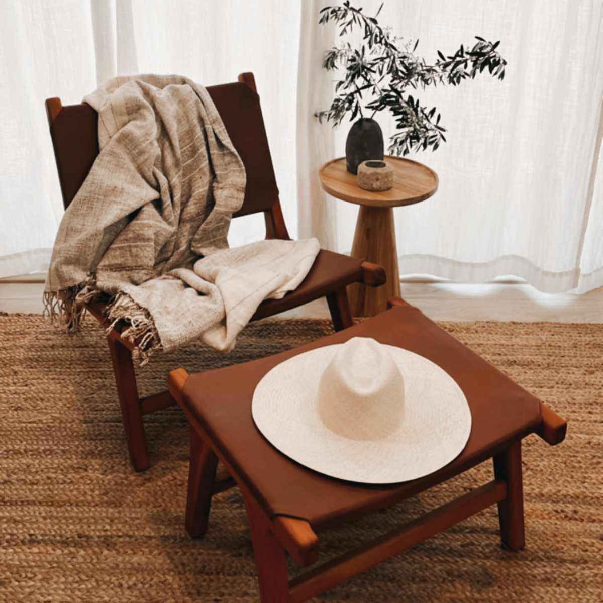 A wooden chair with a blanket draped over it sits beside a small round table, which holds a decorative object and a potted plant, all set against a backdrop of sheer white curtains.