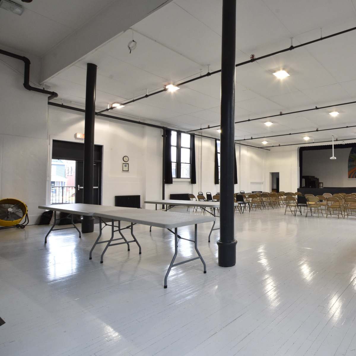An empty community hall with rows of chairs facing a stage, flanked by tables and a bulletin board on one wall.