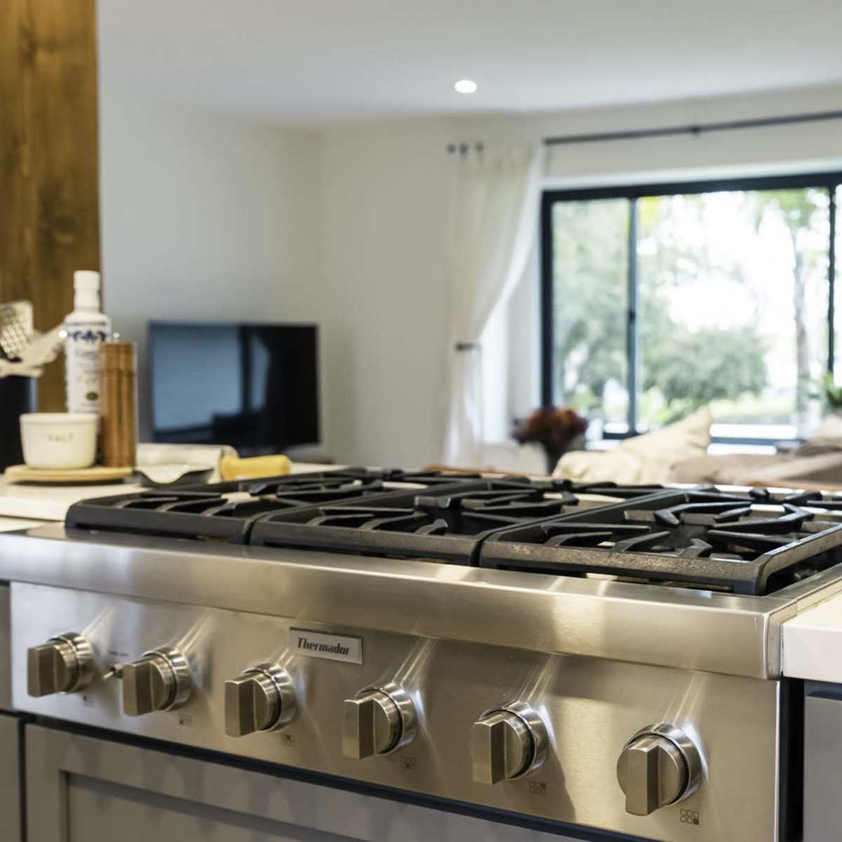 A modern kitchen countertop with a stainless steel gas stove in the foreground and a bright living area visible through a large window in the background.