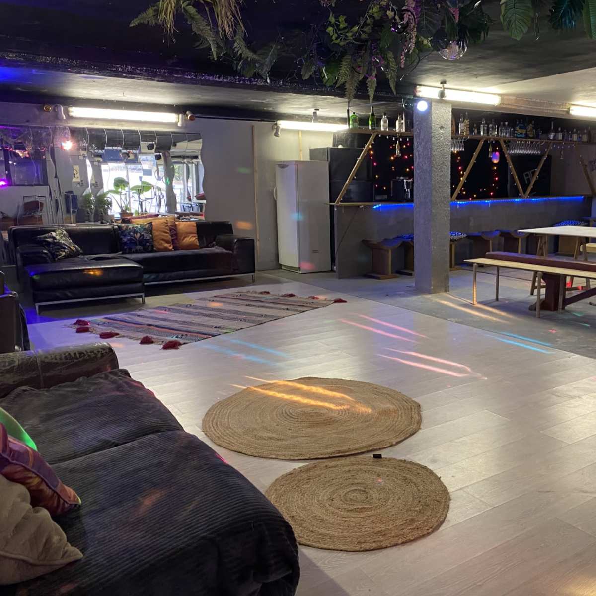 A spacious indoor area with a lounge section featuring sofas, a bar with shelves of bottles, and a table setup, illuminated by colorful lights.