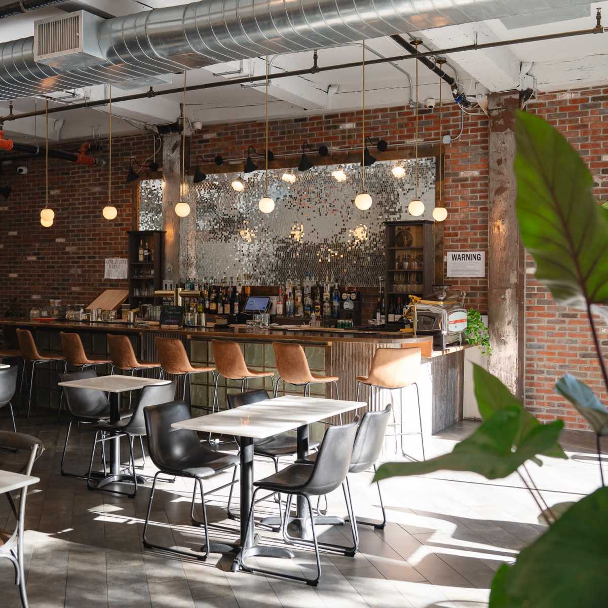 A modern café with brick walls, a bar area lined with bottles, and tables with chairs arranged around the space, illuminated by pendant lights.
