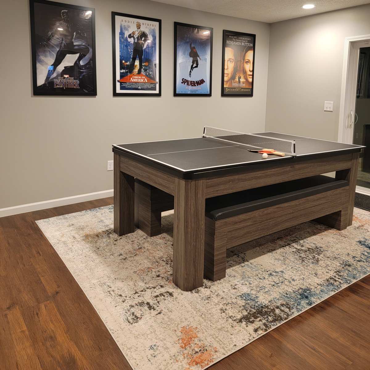A ping pong table is centered in a room with light-colored walls and framed movie posters hanging above it.