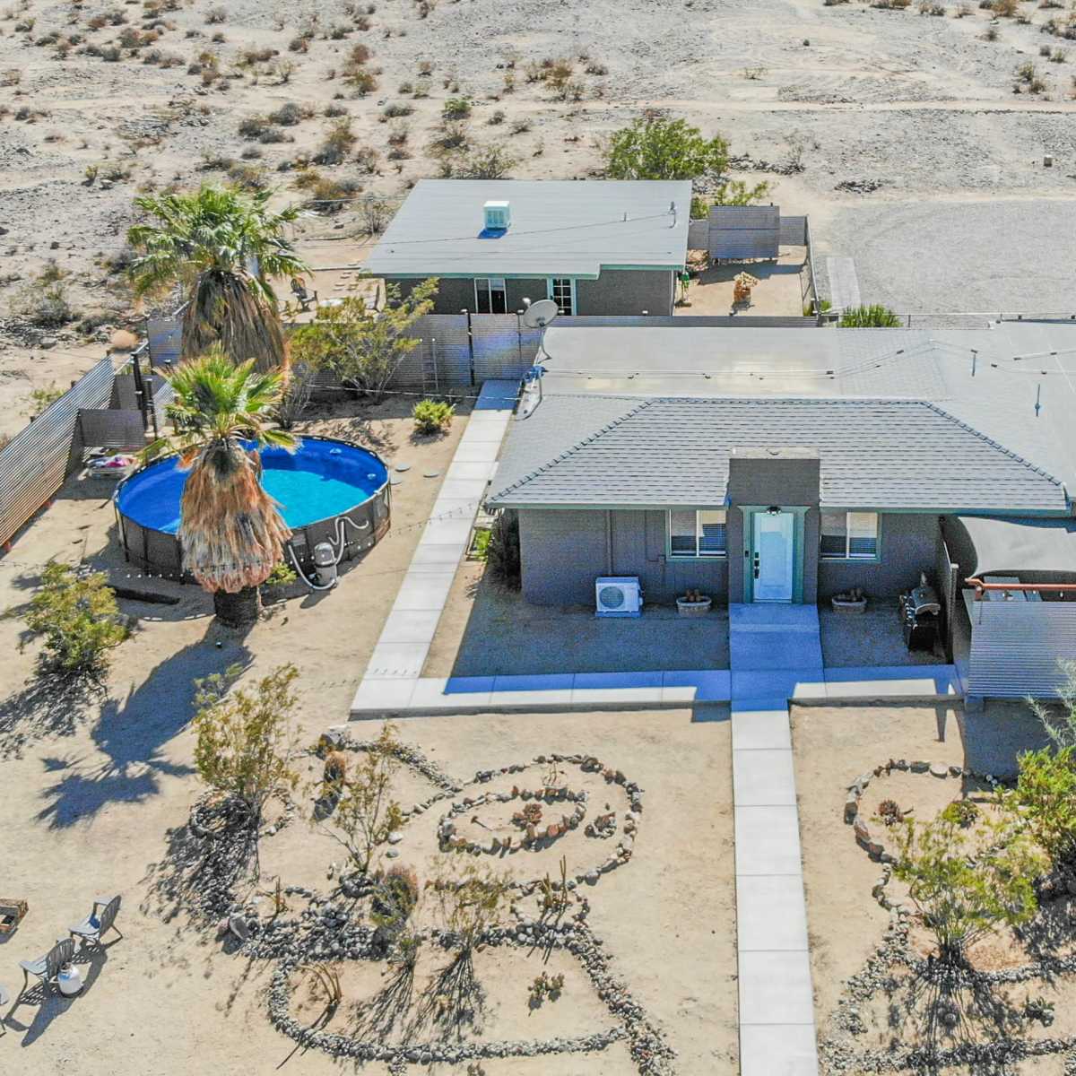 An aerial view of a modest home surrounded by desert landscaping, with a swimming pool, two additional structures, and a neatly laid pathway.