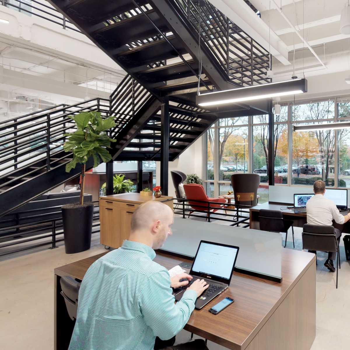 Two people work on laptops at desks in a modern office space with large windows and a spiral staircase.
