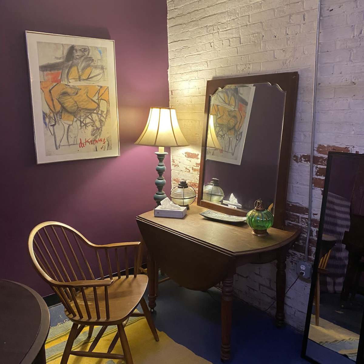 A corner of a room featuring a wooden chair, a dark wooden vanity with a mirror, a lamp, and a colorful rug against a purple wall and exposed brick.