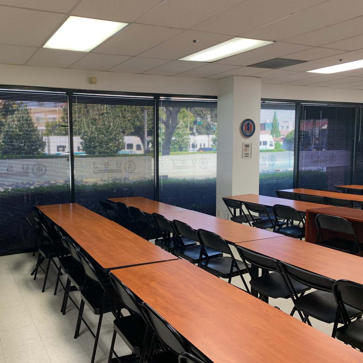 A classroom features long wooden tables arranged in rows with black chairs, and large windows showing a view of a landscaped outdoor area.