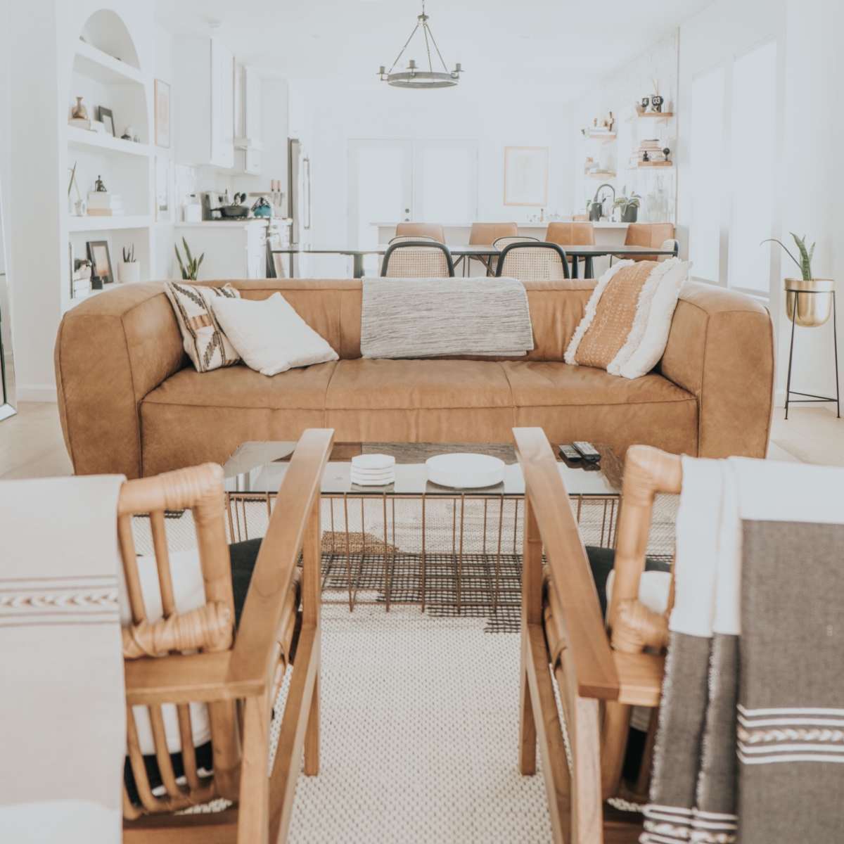 A modern living room features a large brown leather sofa, a coffee table with decorative items, and two wicker chairs, all arranged around a light-colored rug.