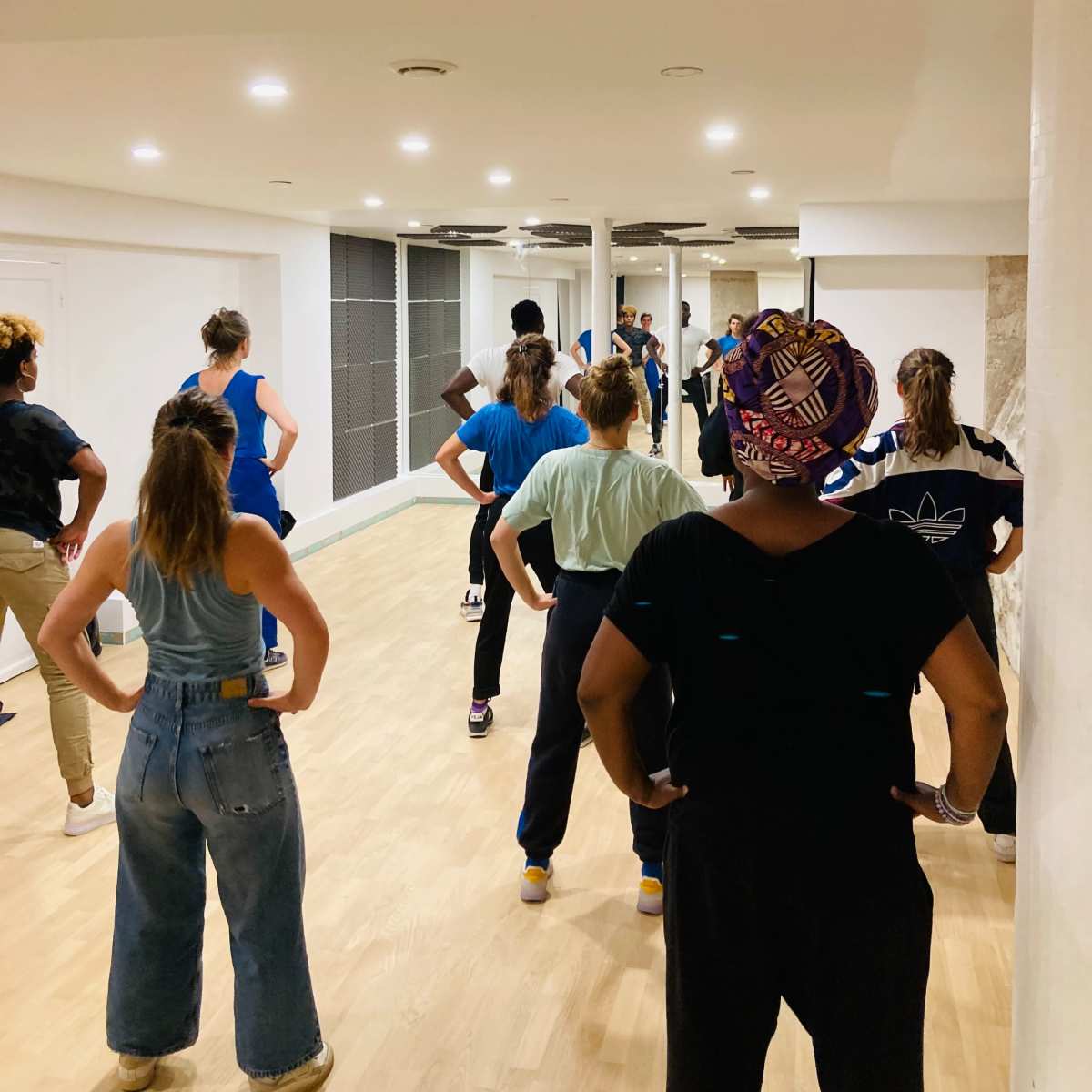 A group of people is practicing dance in a studio with wooden flooring and bright overhead lighting.
