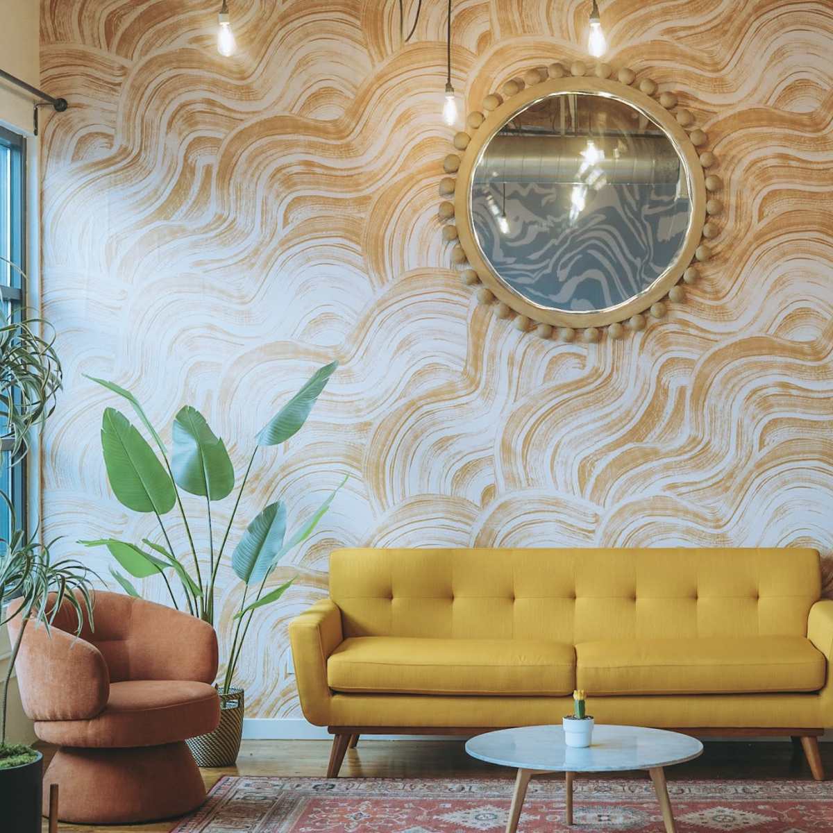 A modern living room featuring a yellow sofa, an orange armchair, a circular mirror, and plants against a wavy patterned wall.