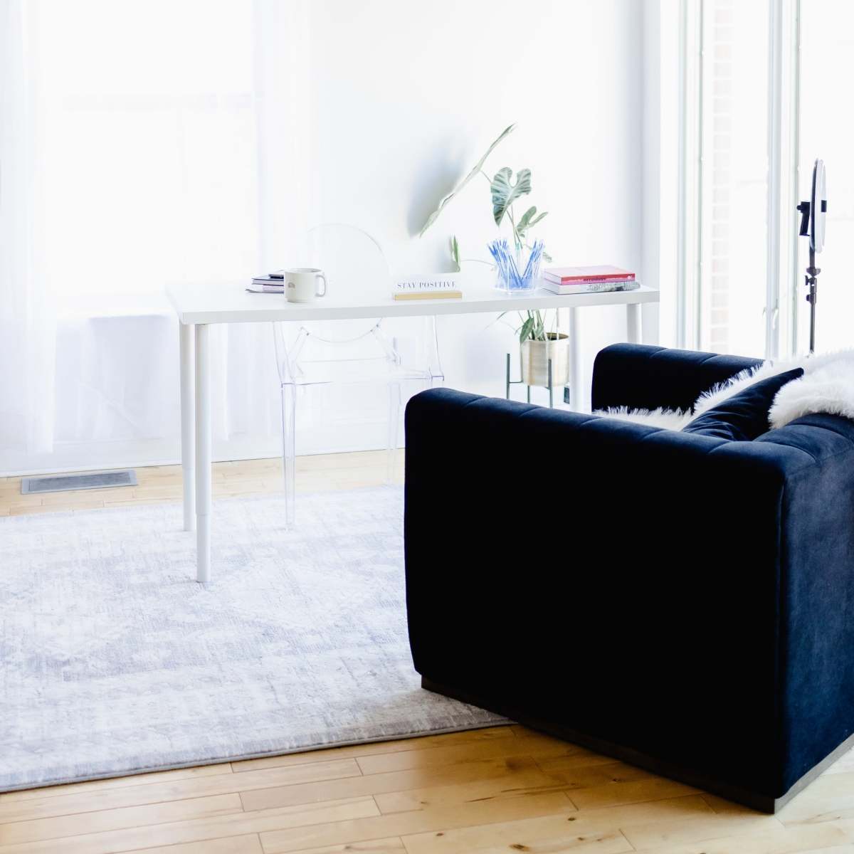 A bright, airy room features a white table and clear chair beside a large window, with a black couch and a light rug on wooden flooring.