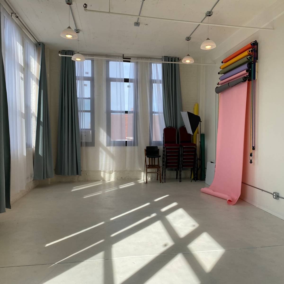 A spacious, well-lit room features large windows, draped curtains, stacked chairs, and a roll of colored backdrop paper against the wall.