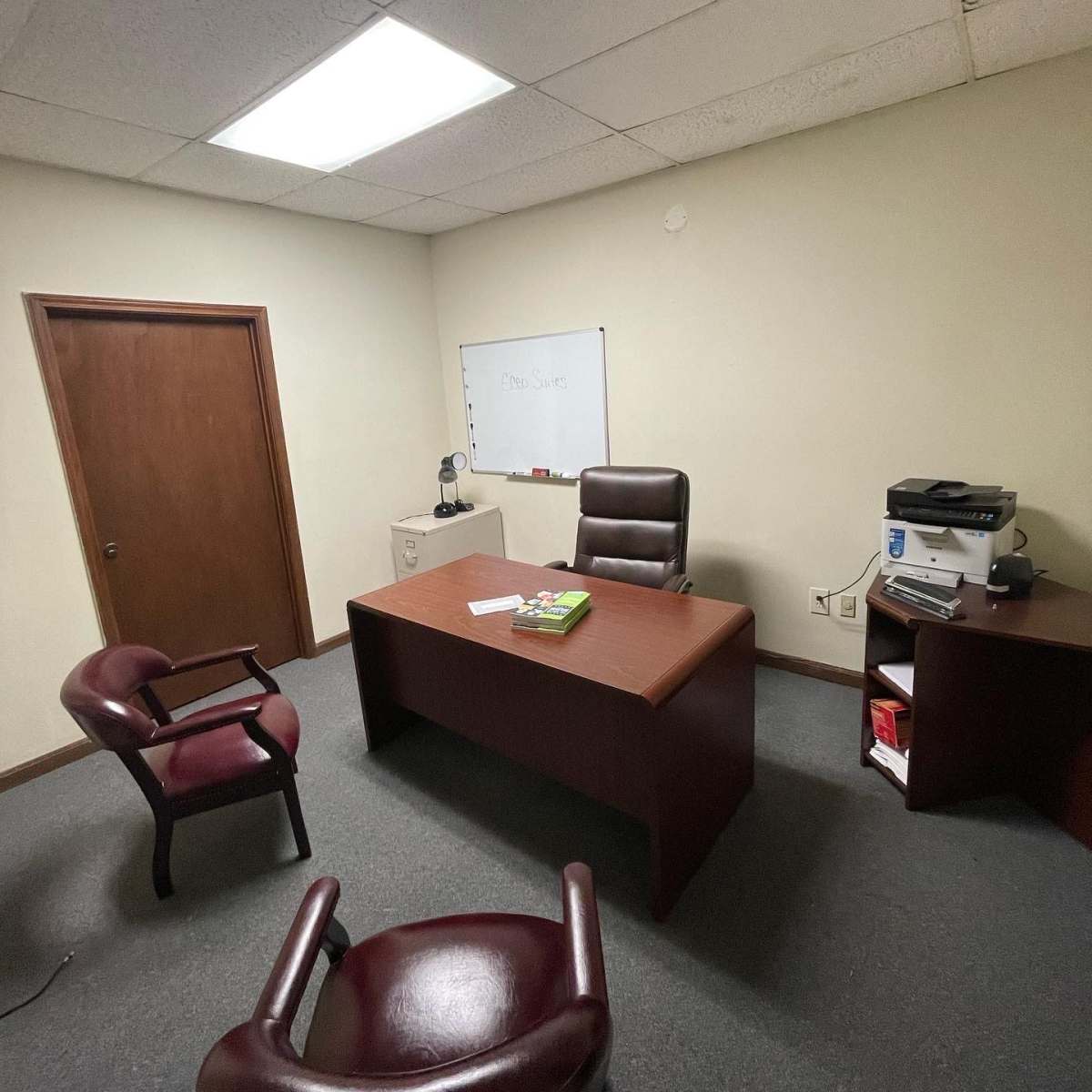A sparsely furnished office with a wooden desk, an office chair, two chairs in front, a whiteboard on the wall, and a printer on a side table.