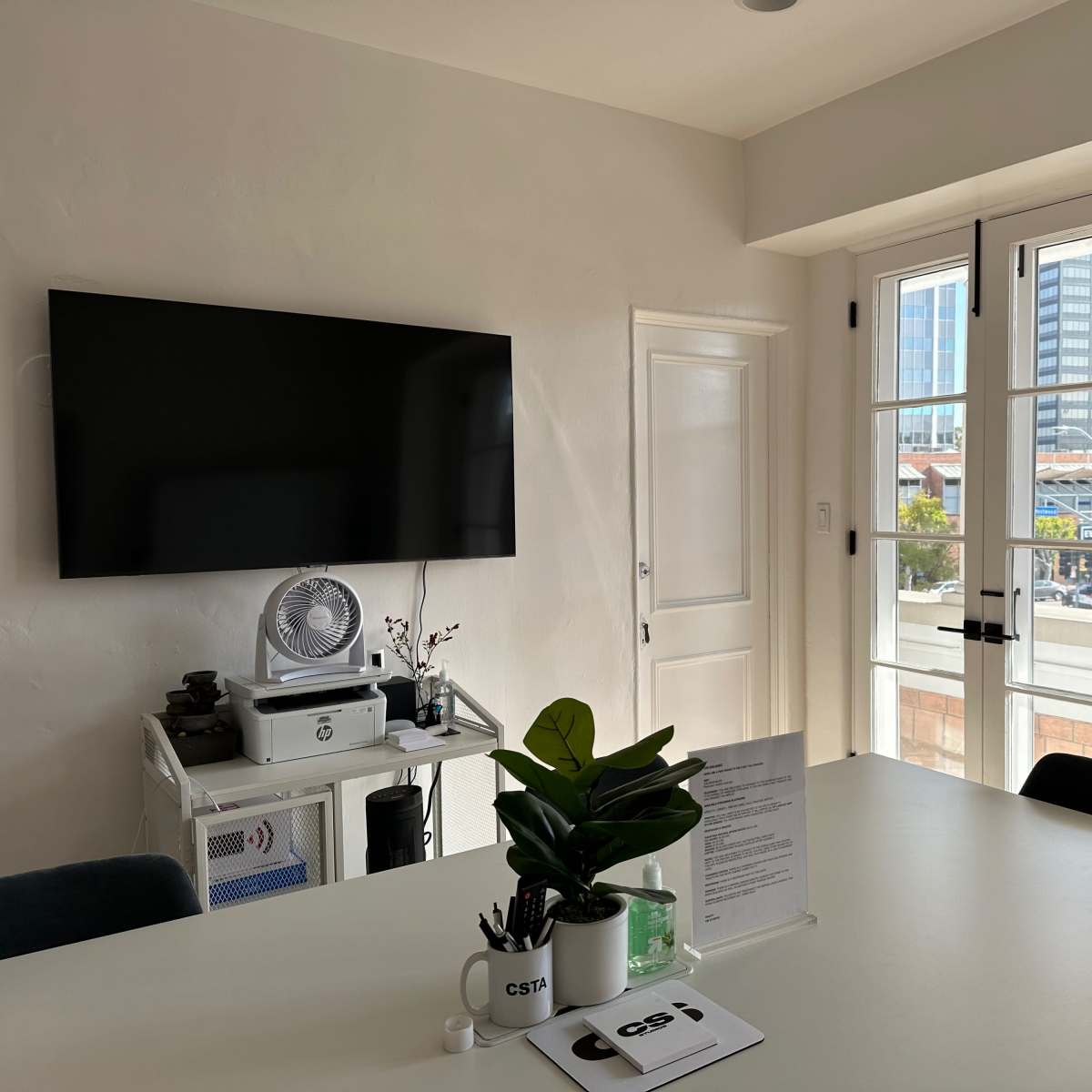 A modern workspace with a large TV mounted on the wall, a table with a plant, and a doorway leading to an outdoor view.