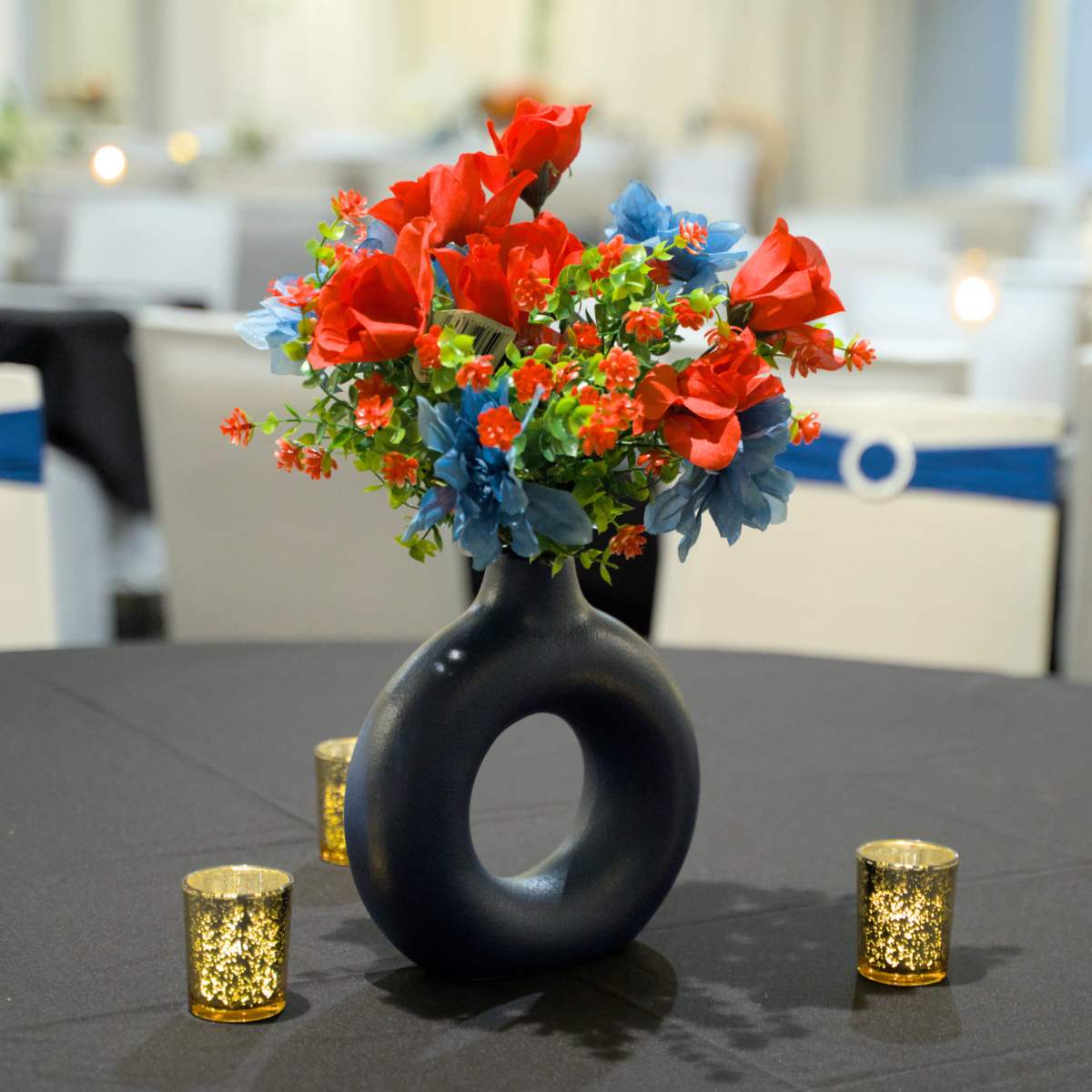 A black table covered with a cloth features a circular vase filled with red and blue flowers, surrounded by glittering candle holders.