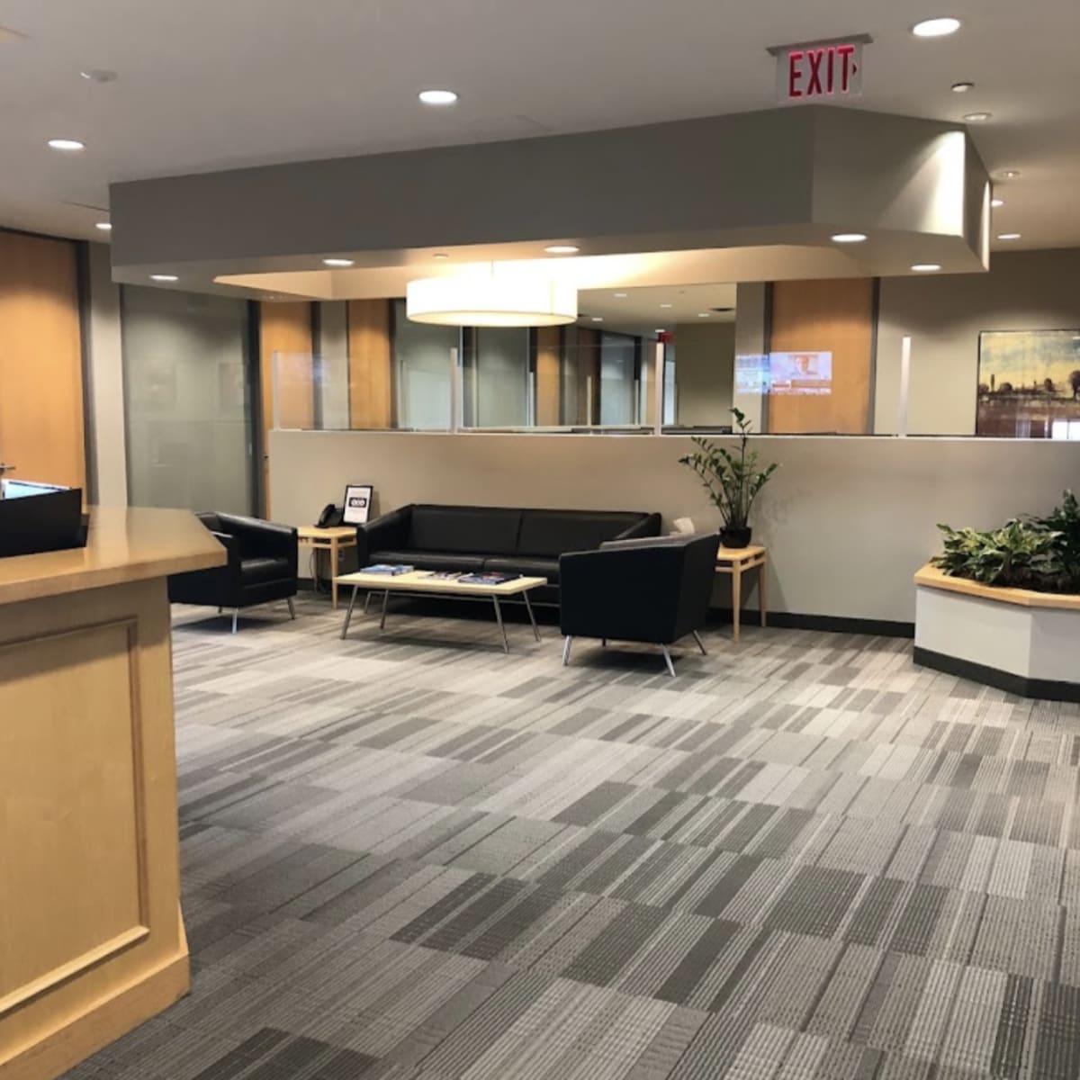 An office reception area features a seating arrangement with a black couch and chairs, along with plants and a large window in the background.