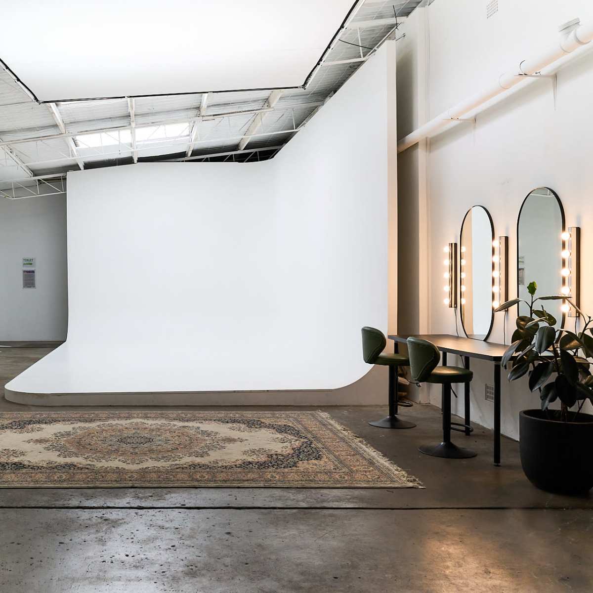 A spacious photography studio with a seamless white backdrop, a large area rug, and a makeup station featuring two mirrors and green chairs.