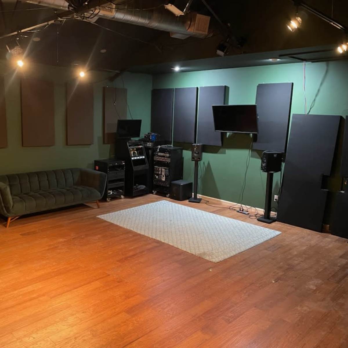 A sound studio with a green wall, wooden floor, a dark sofa, audio equipment, and soundproof panels.
