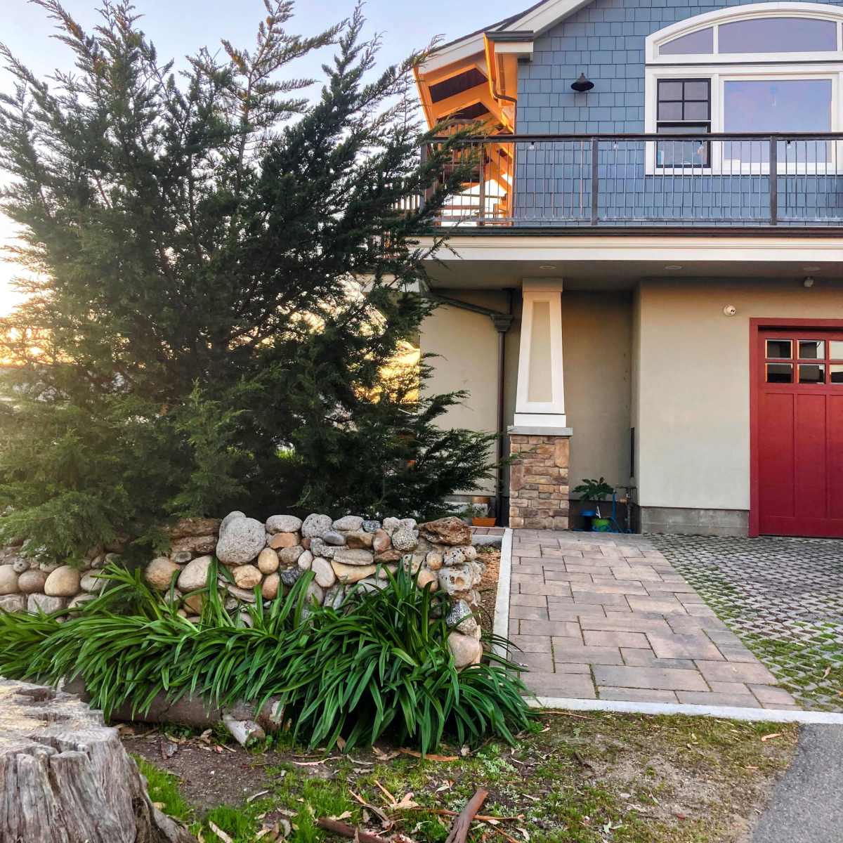 A house with a blue exterior, stone accents, and a red door is surrounded by landscaped greenery and a stone wall.