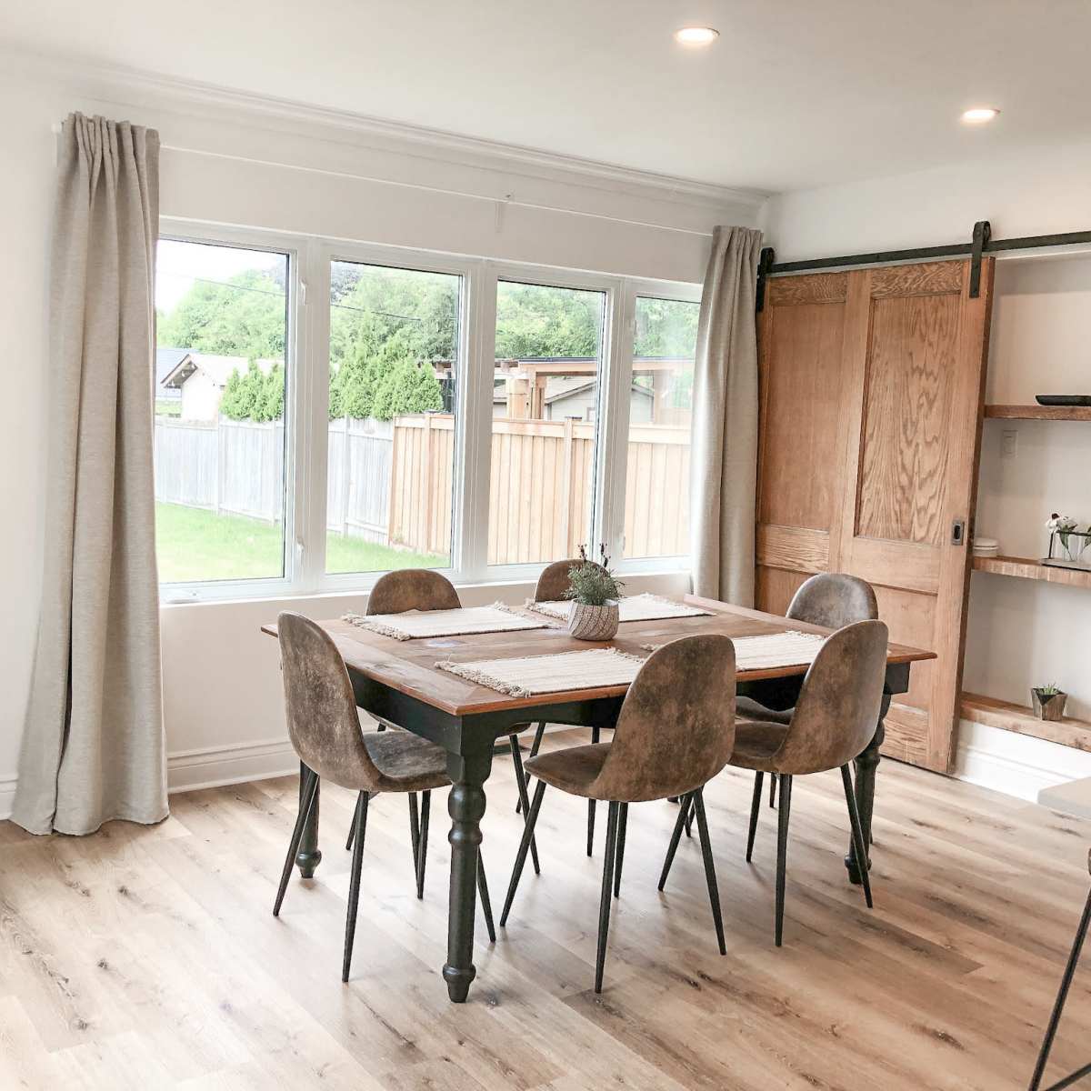 A dining room features a wooden table surrounded by chairs, with a large window and sliding door providing natural light and views of a backyard.