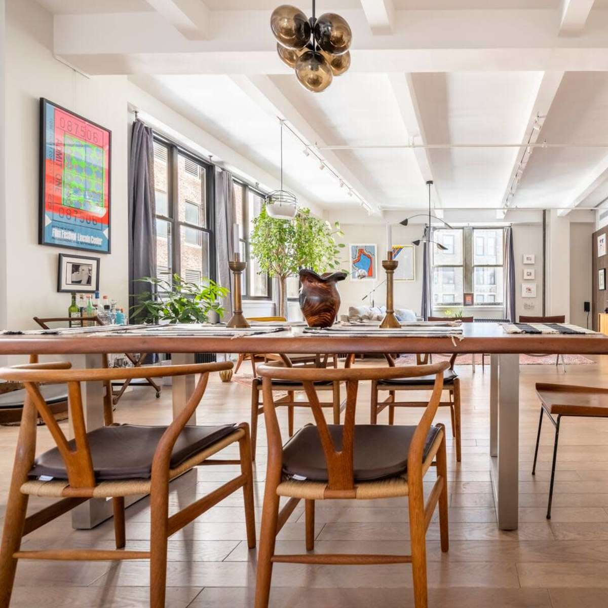 A modern dining area features a long wooden table surrounded by chairs, with art-lined walls and large windows allowing natural light.