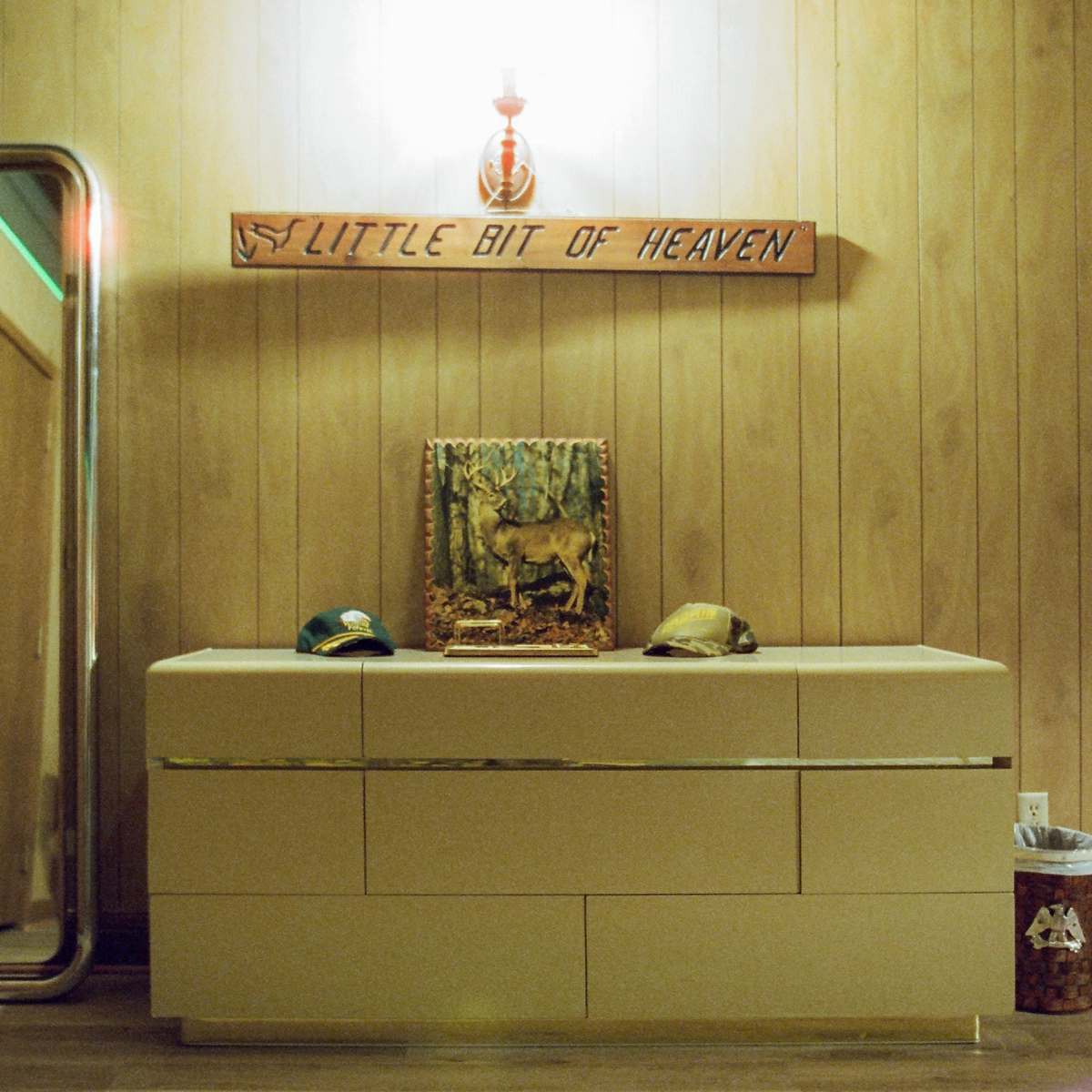 A wooden dresser with two hats and a framed picture of a deer is positioned beneath a wall-mounted light in a room with paneled wood walls.