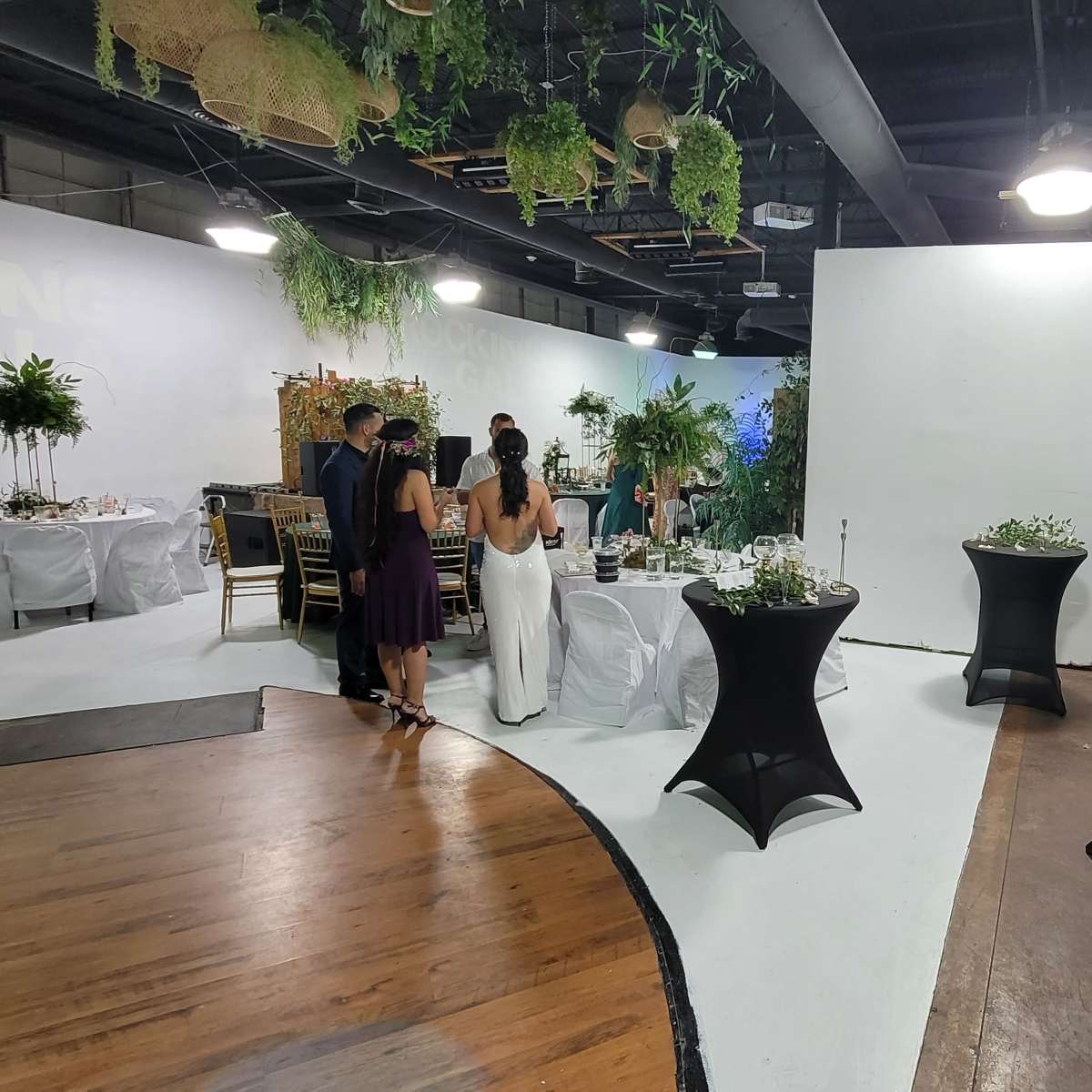 A spacious event venue with tables set for a gathering, featuring white and black decor elements and greenery accents.