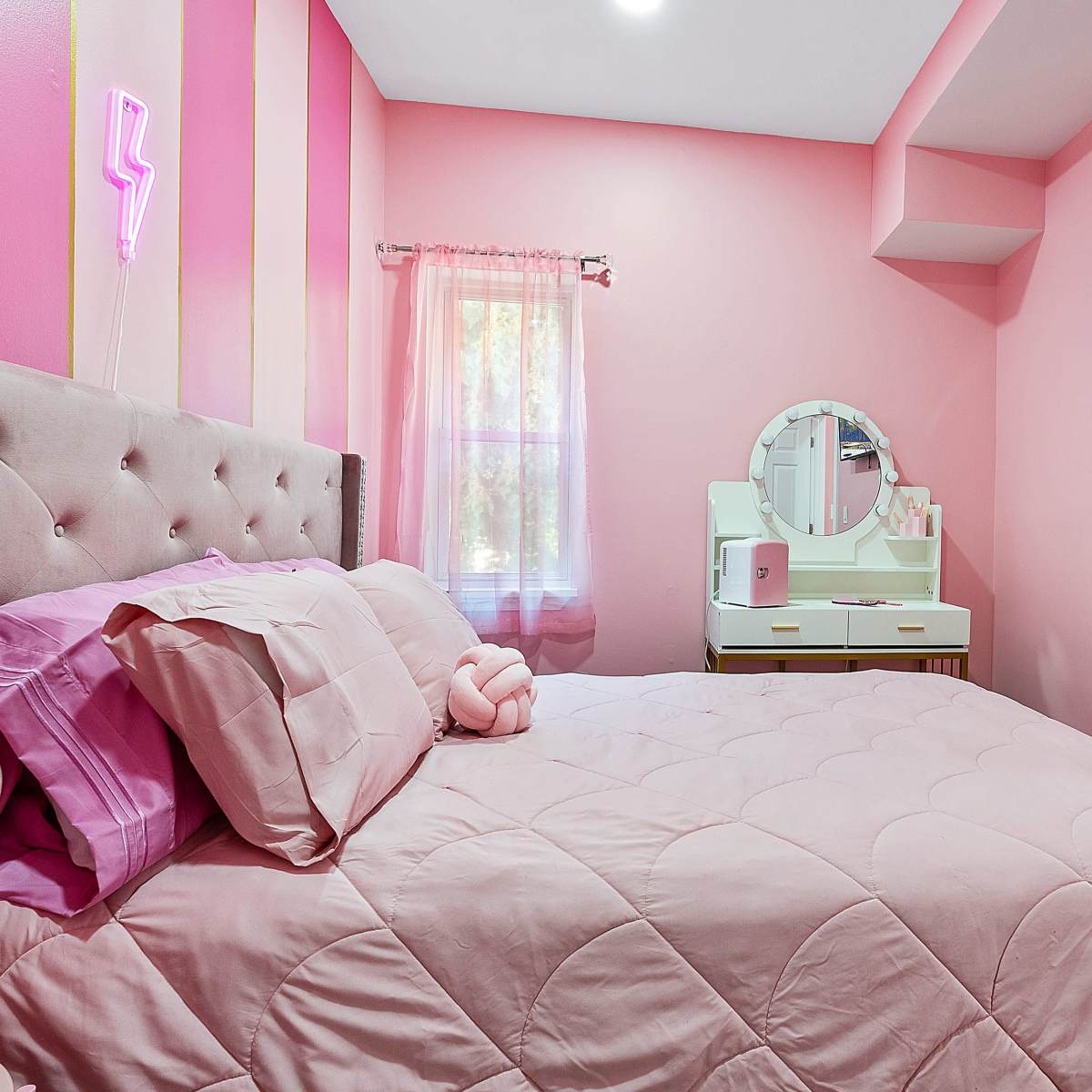 A pink-themed bedroom with a bed, a vanity with a circular mirror, and a television mounted on the wall.