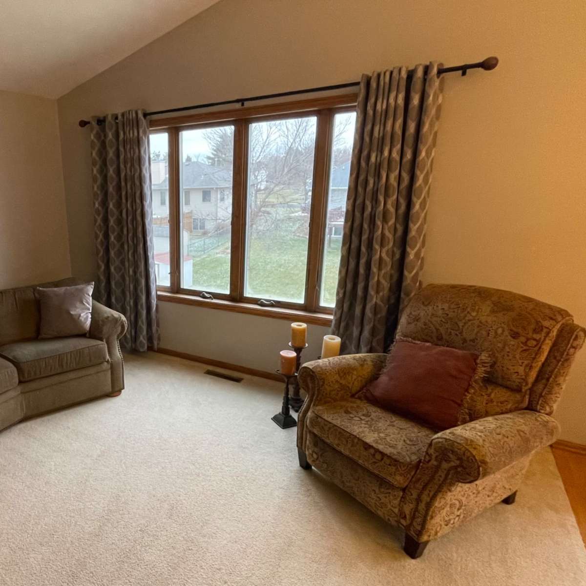 A cozy living room features a curved sofa, an armchair, and large windows with drapes overlooking a yard.