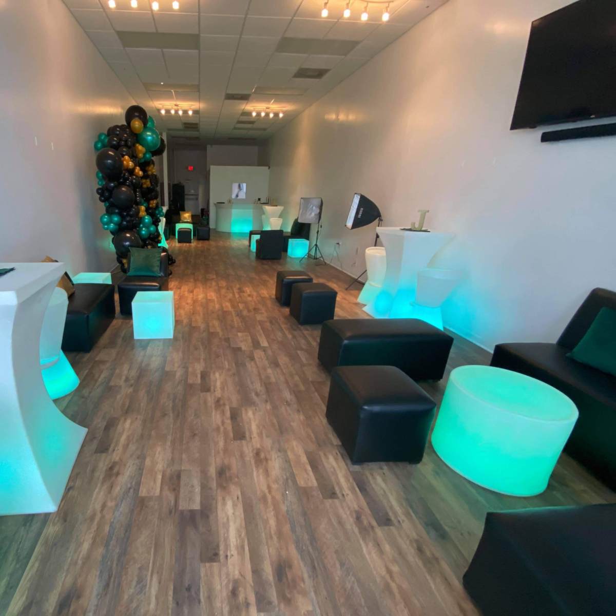 A spacious indoor venue featuring a long, wooden floor, black seating, illuminated green and white tables, and a decorative balloon display in one corner.