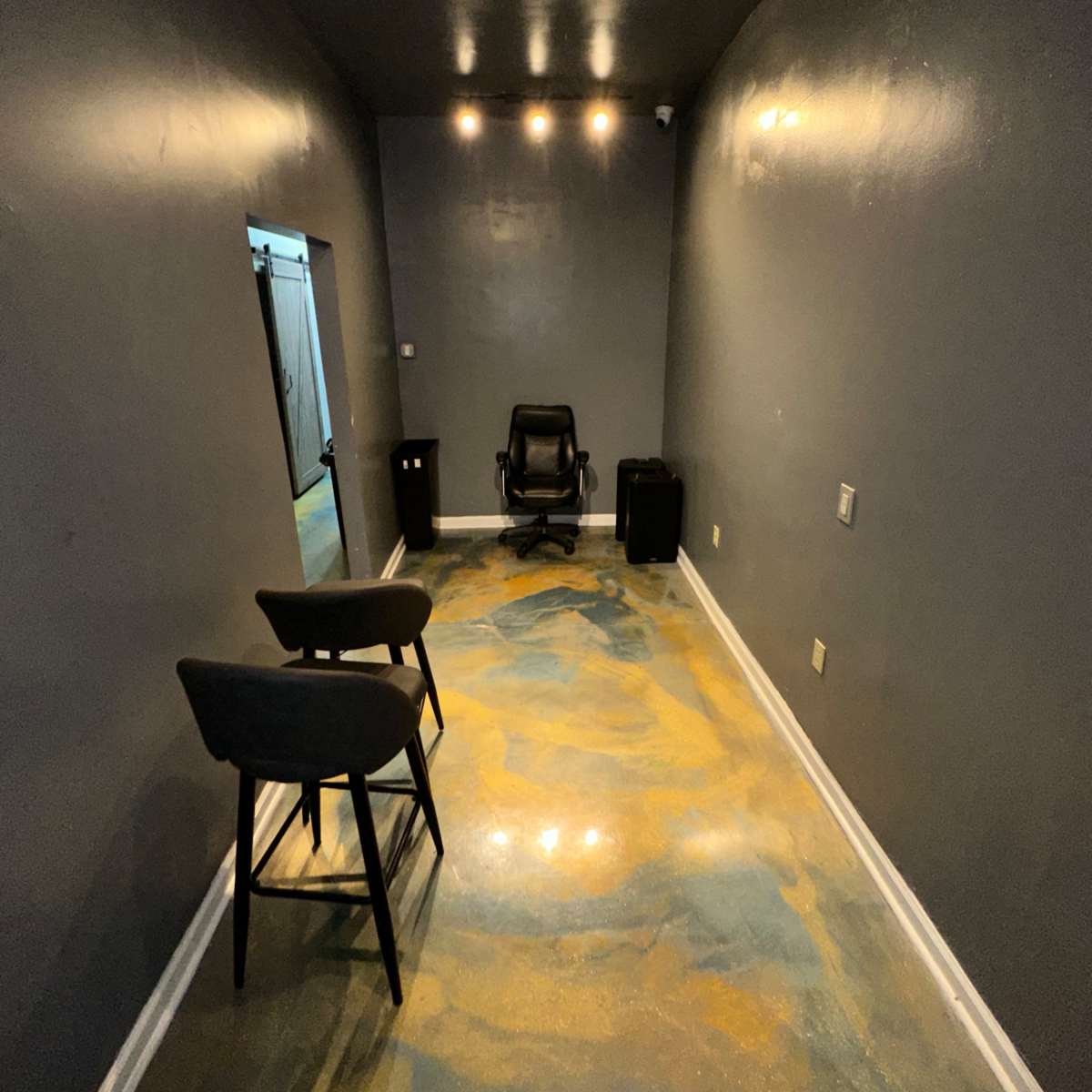 A narrow, empty room with gray walls, a shiny floor featuring blue and gold designs, two stools, an office chair, and a mirror on one side.