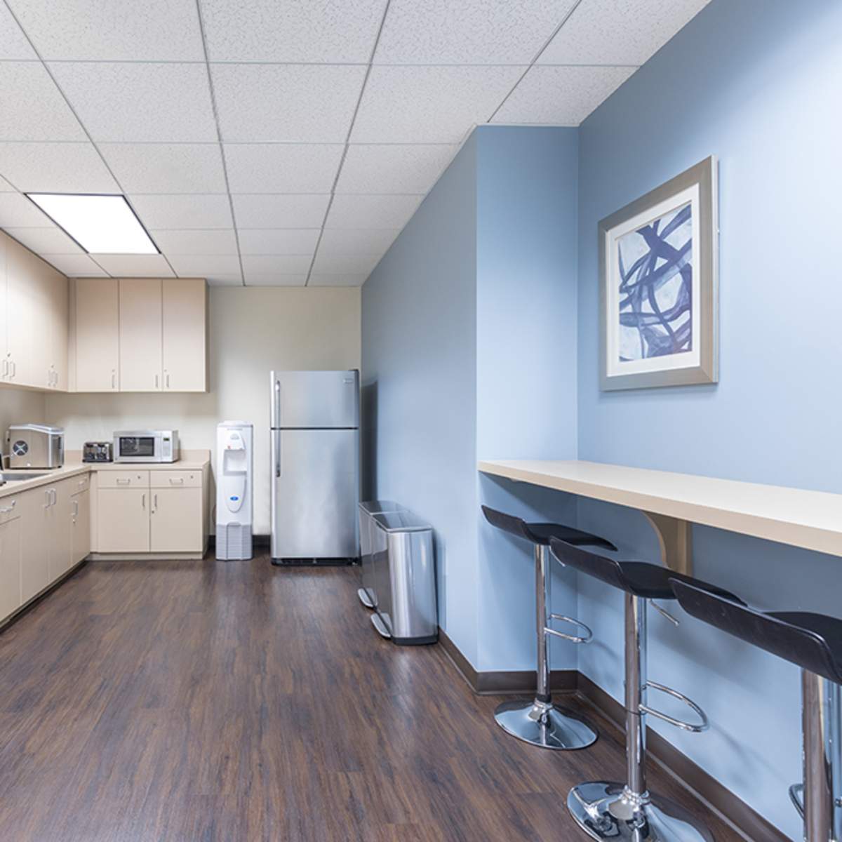 A break room with beige cabinets, a countertop, a refrigerator, a microwave, and a coffee machine against a blue wall.