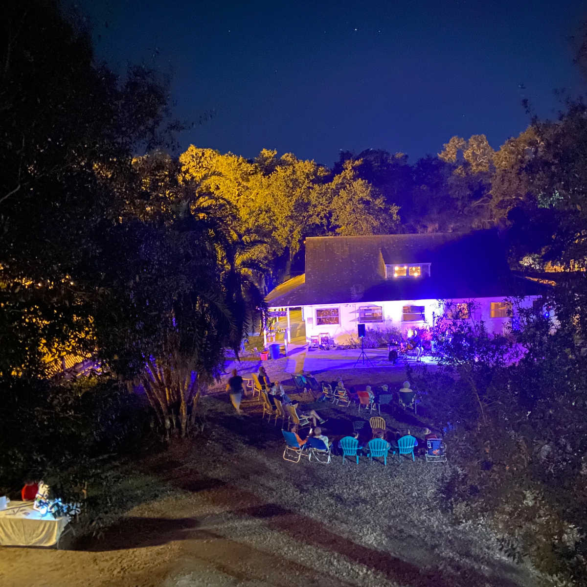 A house is illuminated by colorful lights at night, surrounded by chairs arranged on a grassy area amidst trees.