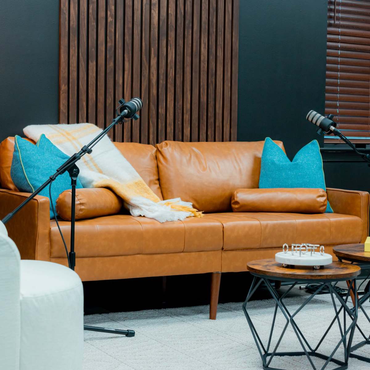 A cozy seating arrangement with a brown leather sofa adorned with turquoise pillows and a white armchair, accompanied by microphones on stands and a wooden coffee table.