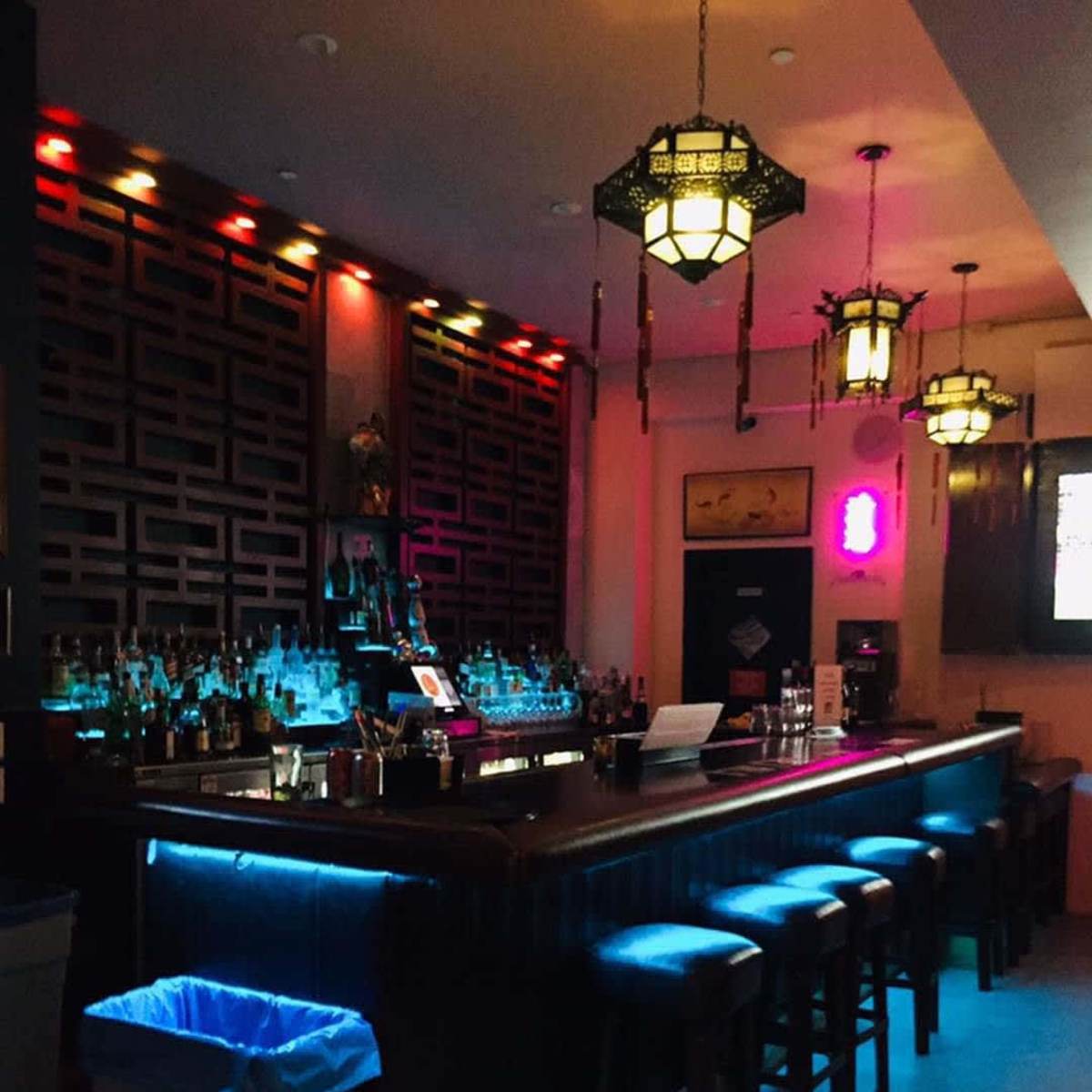 A dimly lit bar features a long wooden counter with several stools and decorative lanterns overhead.